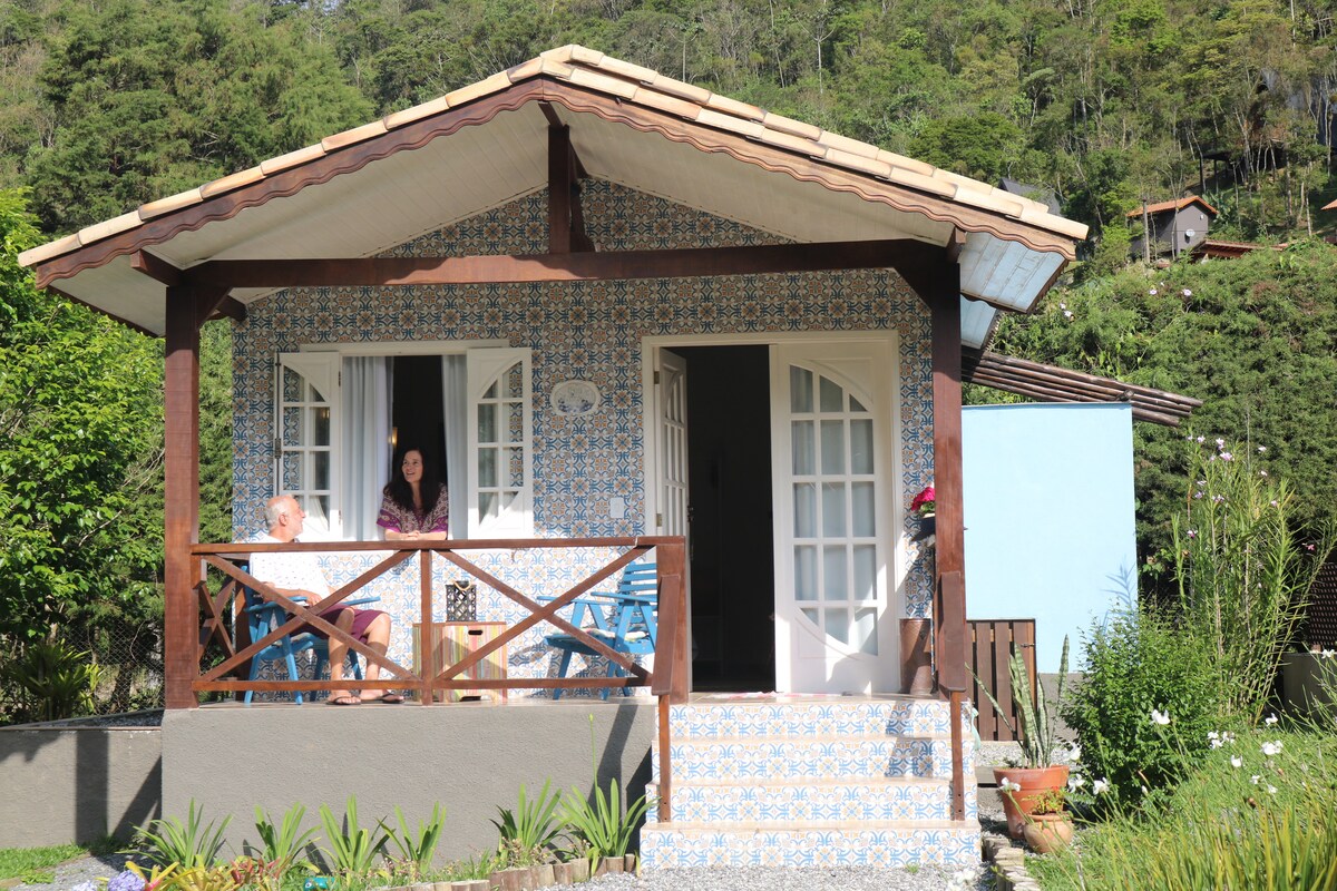 A charming chalet with a tiled facade is shown, featuring a traditional sloped roof. Two white doors and two windows frame the entrance. A seating area, furnished with blue chairs, is positioned on the porch. Lush greenery and flowers surround the building, enhancing the outdoor setting.