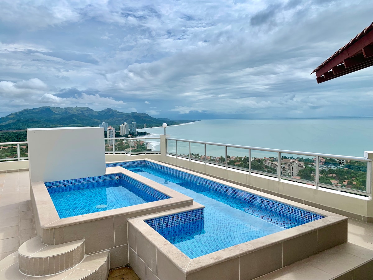 A rooftop area features two inviting pools, one being a spacious hot tub. A panoramic view of the ocean and distant mountains is visible beyond the pools. The deck offers ample space for relaxation, complemented by a railing that enhances safety.