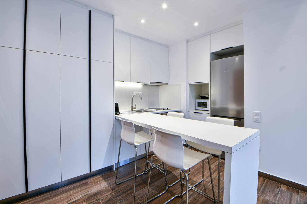A modern open-plan kitchen is featured, characterized by sleek white cabinetry and a minimalist design. A spacious countertop with high stools is positioned in front, while built-in appliances are integrated seamlessly within the cabinetry. Bright lighting illuminates the area, highlighting its clean lines.