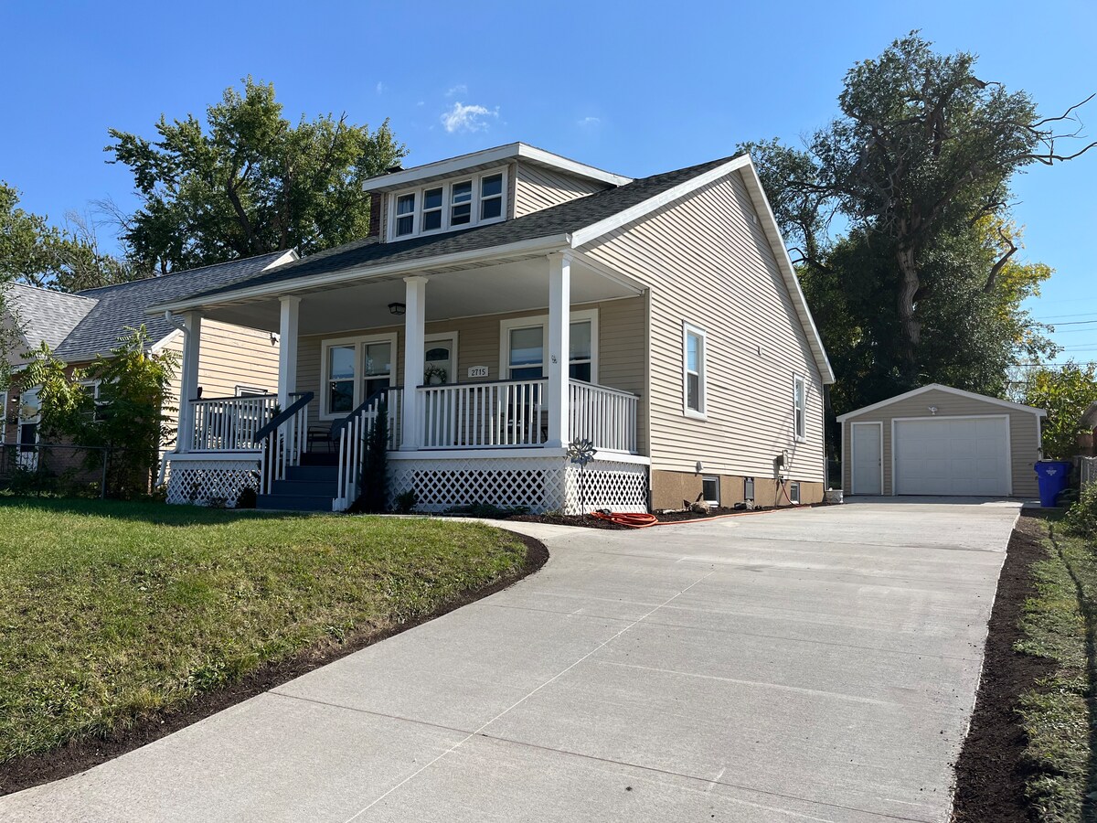 The exterior of the house features a welcoming front porch supported by white columns. A well-maintained lawn and driveway are visible, leading to a one-car garage. Tall trees provide shade, enhancing the home's inviting appearance.