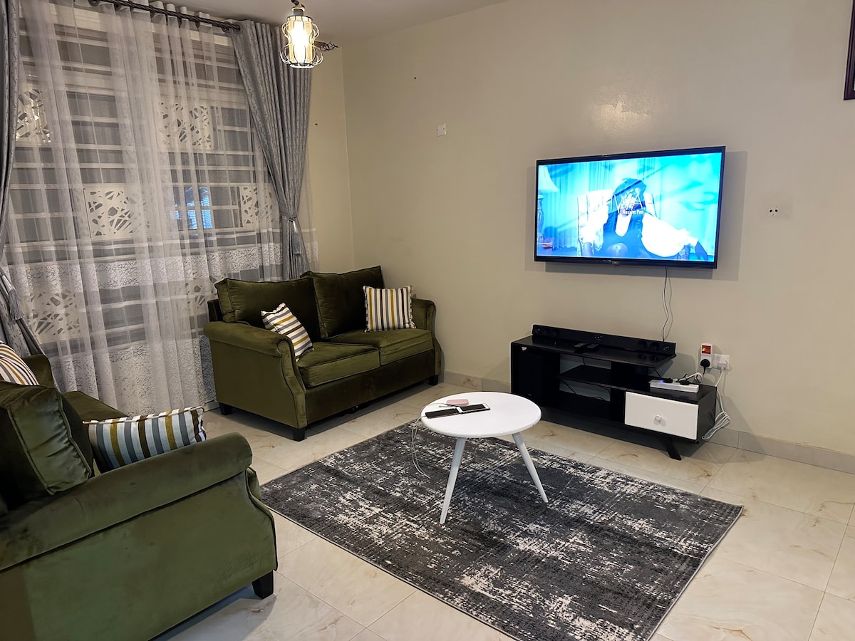 A cozy living area features two green sofas with striped cushions arranged around a white coffee table. A flat-screen TV is mounted on the wall opposite, with a stylish media console underneath. Natural light filters in through sheer curtains along a large window.