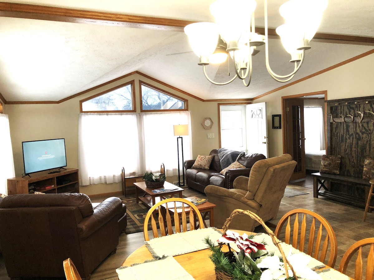 An inviting living area is presented, featuring a mix of comfortable seating, including a large brown sofa and two armchairs. Natural light filters in through large windows, enhancing the overall spaciousness. A dining table with wooden chairs is positioned nearby, adding functionality to the space.