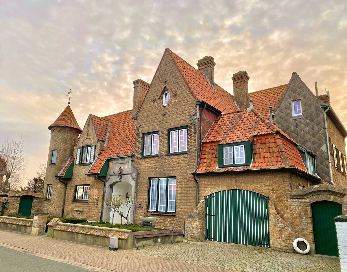 The exterior of a distinctive building features a combination of brick and stone, highlighted by a variety of gabled roofs and vibrant green shutters. A circular tower is complemented by an arched entryway. A cobblestone driveway leads to a green gate, enhancing the architectural charm.