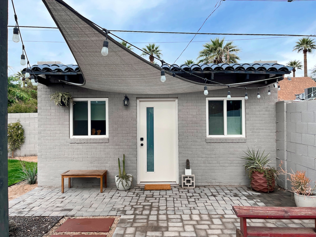 The guesthouse exterior showcases a simple façade with light gray brick walls, a blue tile roof, and large windows. A colorful shade structure provides coverage over the entrance, while string lights enhance the outdoor ambiance. Potted plants and benches create a welcoming entryway.