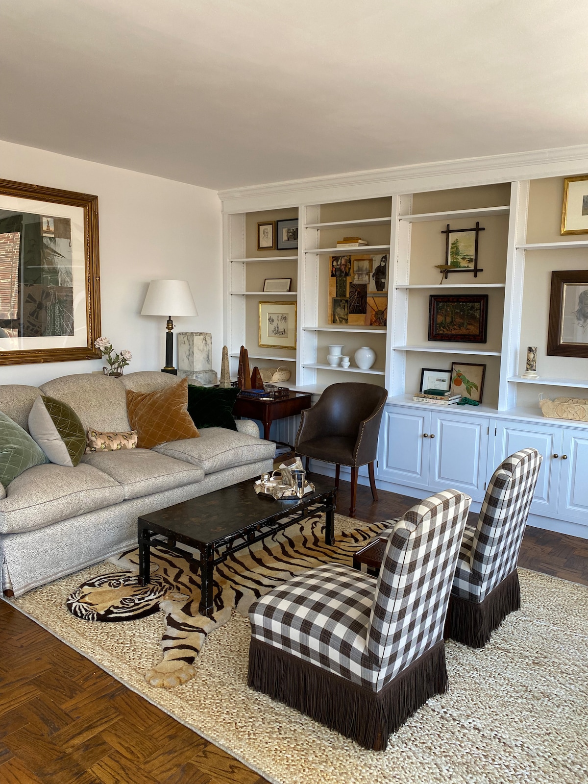 A comfortable living area features a gray sofa adorned with decorative pillows and a patterned throw. Two plaid armchairs flank a black coffee table, which sits on a textured area rug. A wall unit displays framed artwork and decorative items, contributing to a curated ambiance.