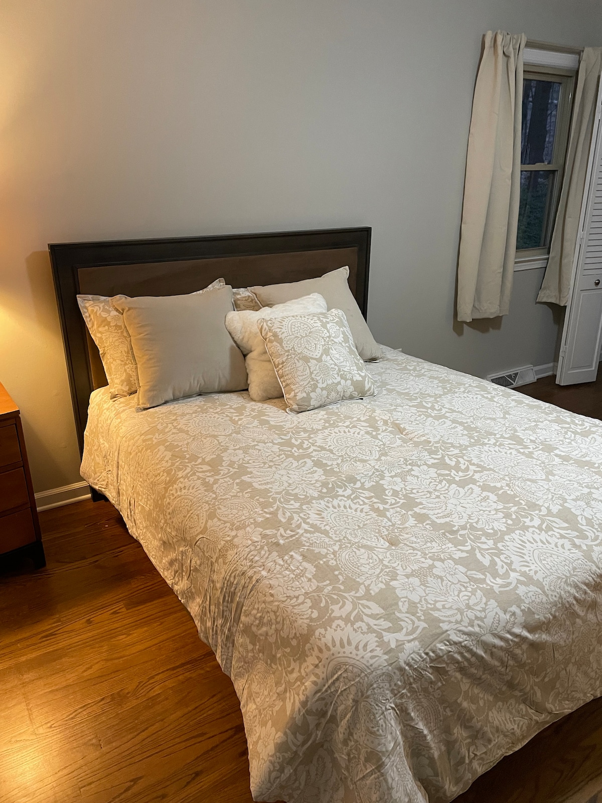 A comfortable sleeping area is presented, featuring a spacious bed adorned with a light-patterned quilt and decorative pillows. Warm wooden flooring and soft lighting from a bedside lamp create a soothing ambiance. A window offers natural light, framed by light-colored curtains.