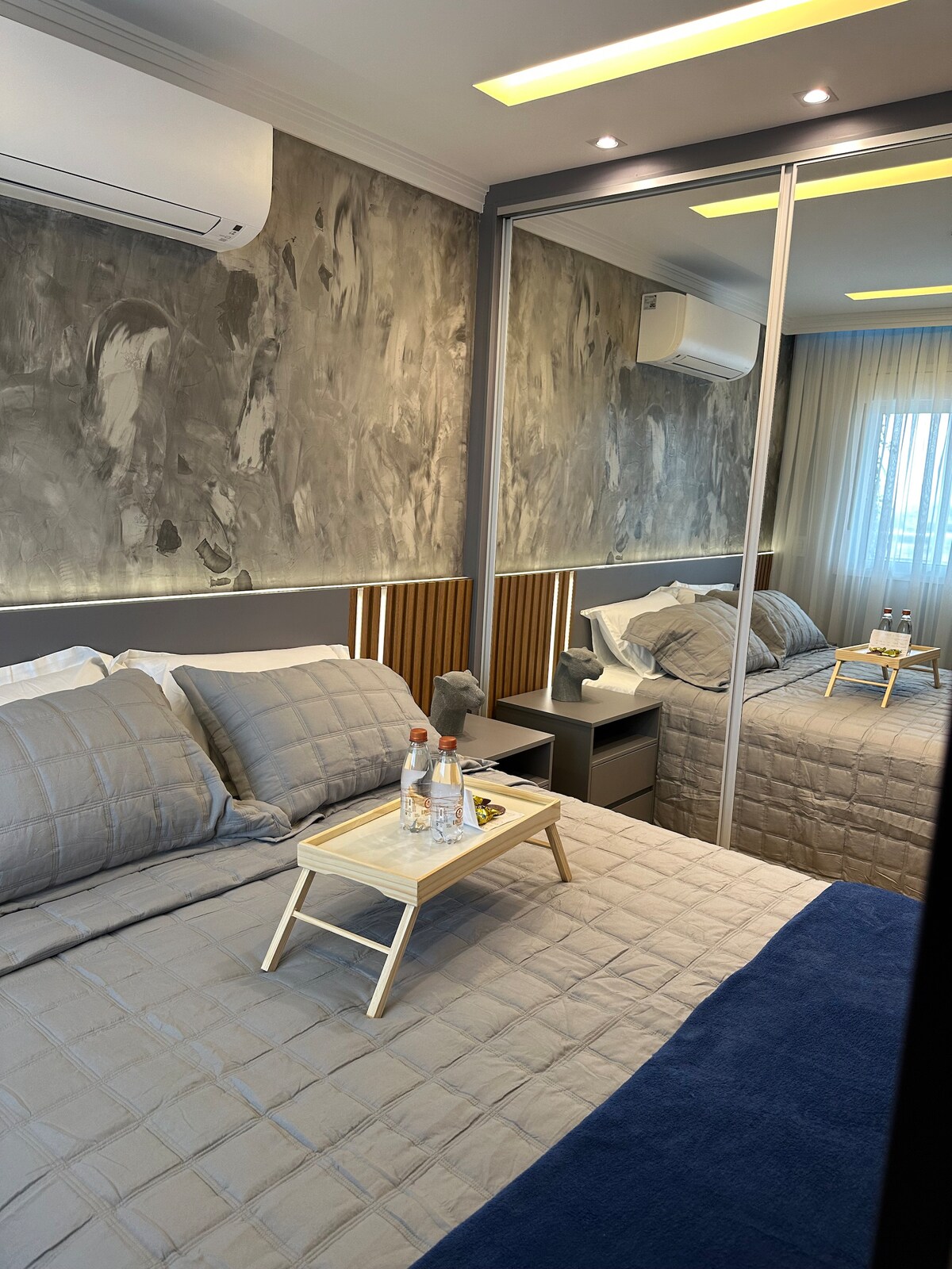 A modern bedroom features a spacious bed with neutral bedding. A mirrored wall reflects natural light, enhancing the room's brightness. A small wooden tray holds two bottles of water and a snack, situated on the bed's edge. Air conditioning is visible above.