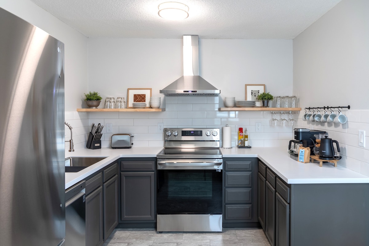 The kitchen features modern appliances including a stainless steel stove and refrigerator. Open shelving holds glassware and decorative items. A coffee maker and toaster are present on the countertop, along with a double sink and ample workspace in a well-organized layout.