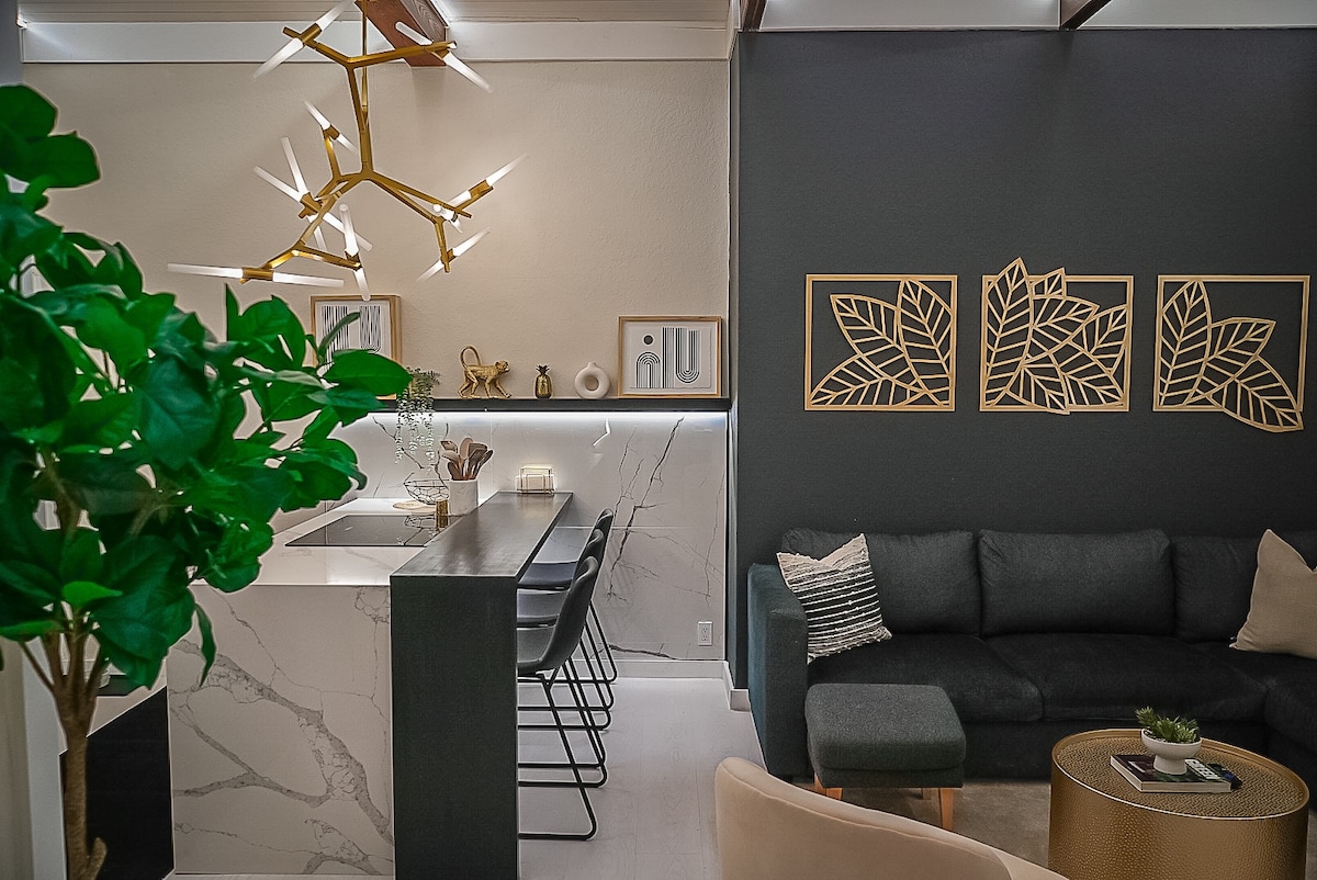 An open concept living area features a modern kitchen island with bar seating and elegant lighting overhead. A dark-colored sofa is complemented by a light-colored accent pillow, and framed wall art adds a touch of style. A large potted plant provides a hint of greenery.