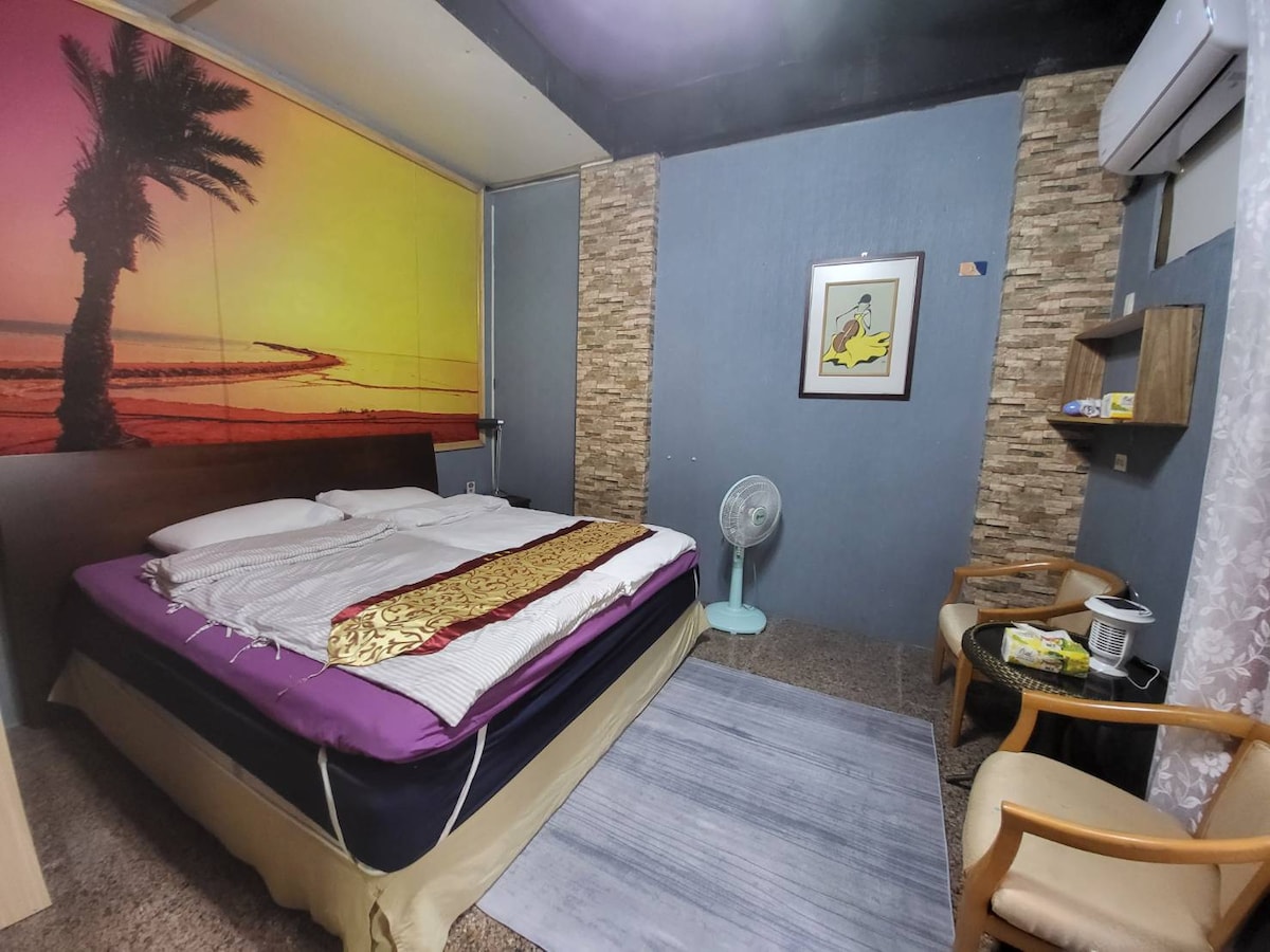 A spacious sleeping area features a large bed with a decorative blanket, set against a backdrop of a sunset beach mural. Natural light filters through a nearby window, complemented by a fan and two wooden chairs placed beside a small table.