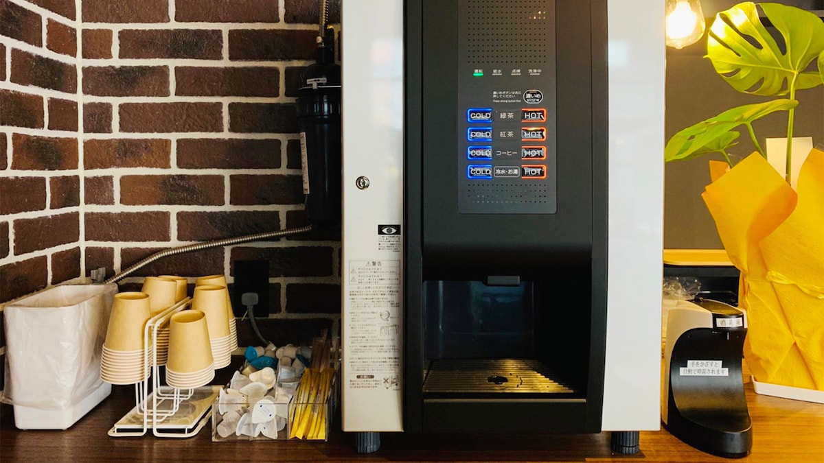 A coffee machine is positioned on a wooden counter, featuring a variety of beverage options displayed on its front panel. Disposable cups are neatly stacked beside the machine, accompanied by straws and sugar packets. The backdrop is accented by a brick pattern, enhancing the overall aesthetic.