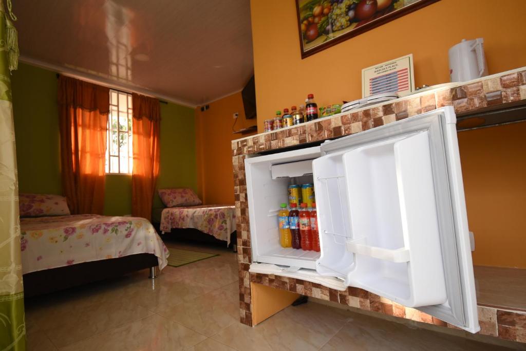 A compact kitchenette is visible with an open refrigerator stocked with beverages. In the background, two cozy beds adorned with floral bedding are located against the wall. Natural light filters through a nearby window, illuminating the colorful room with vibrant walls.