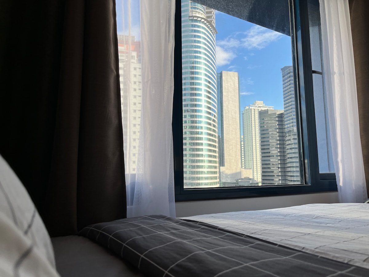 A view from a cozy sleeping area features large windows overlooking a cityscape filled with tall buildings and a bright blue sky. Sheer curtains allow natural light to illuminate the space, creating a serene atmosphere.