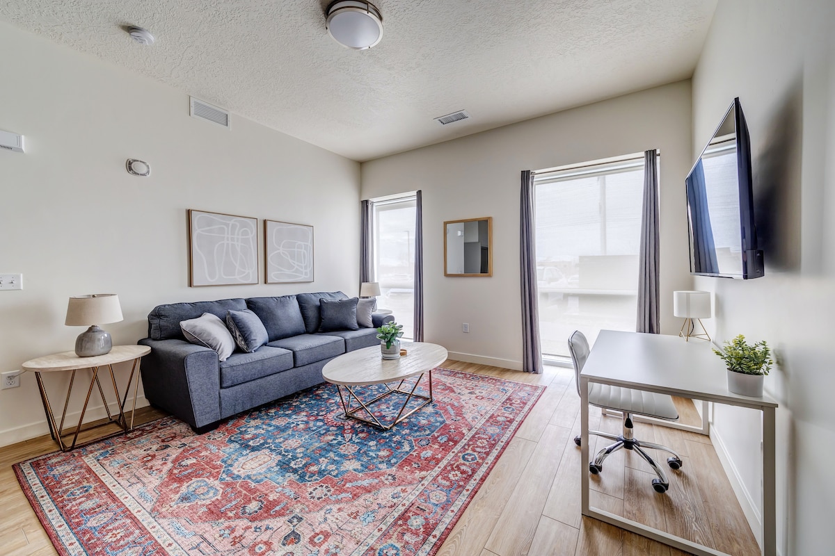 A modern living space features a comfortable gray sofa positioned next to a decorative area rug. A sleek coffee table rests in front, and a wall-mounted television is visible. Natural light enters through two large windows, complementing a designated workspace with a desk and chair.