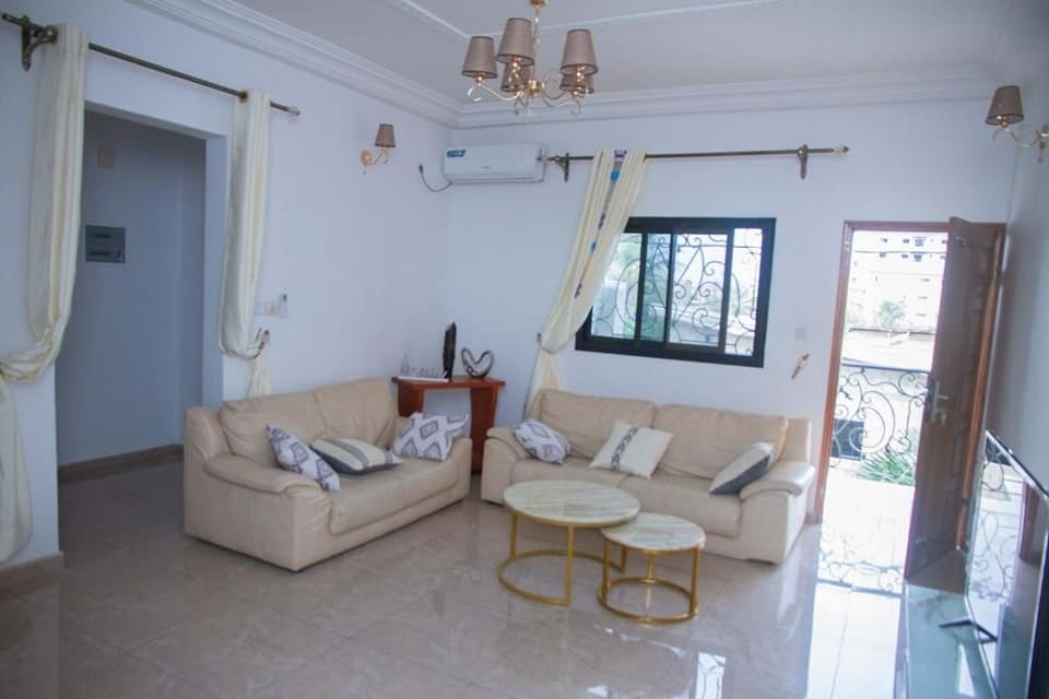A light-filled living area showcases two cream-colored sofas arranged around a circular coffee table. Elegant curtains frame the window, while the air conditioning unit is mounted above. A decorative side table complements the space, and an open door leads to a balcony.