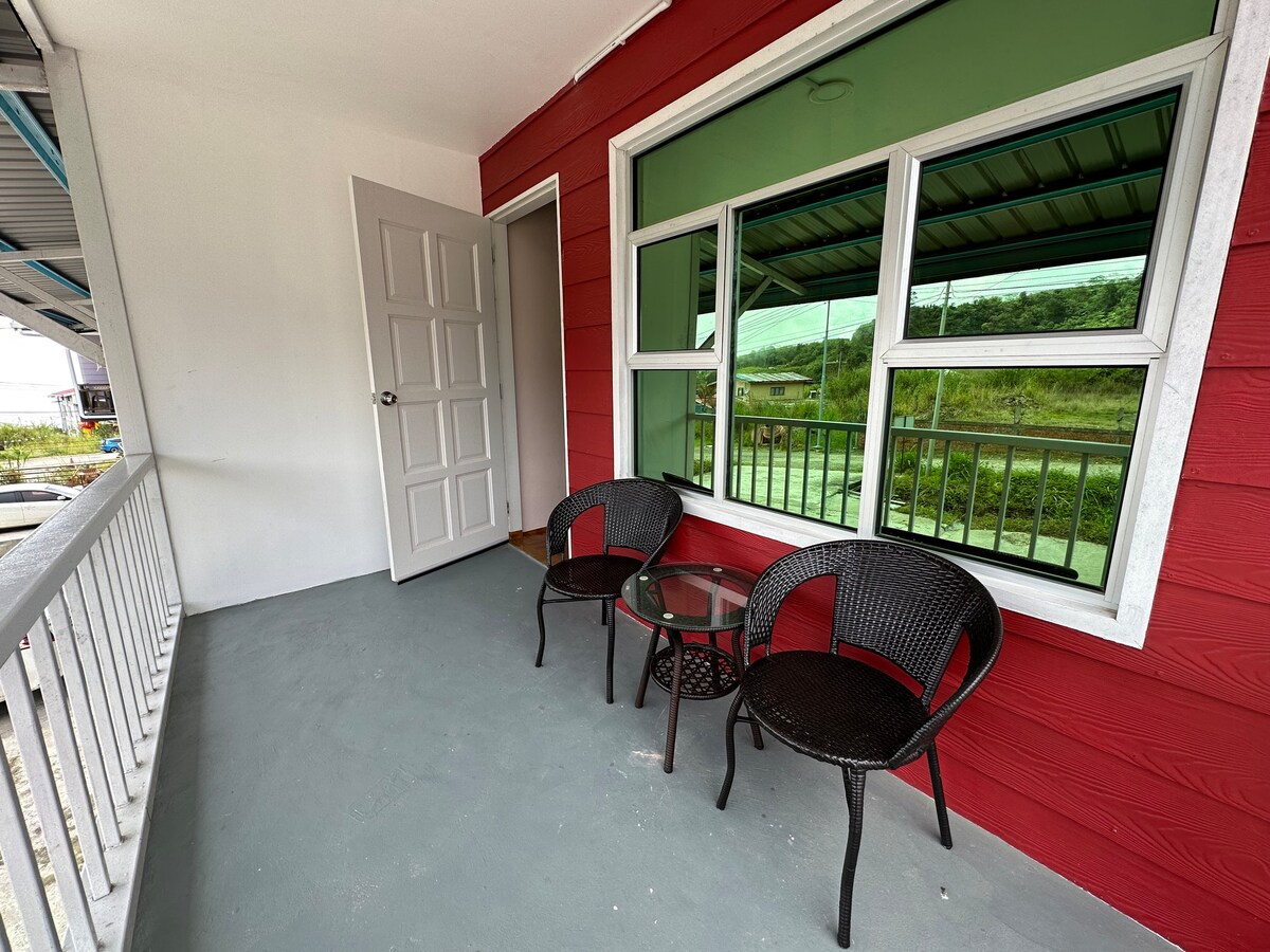 A covered porch features a red exterior wall and large windows providing views of the surroundings. Two wicker chairs and a small glass table are arranged on the floor, creating a space for relaxation. A door leads into the interior of the accommodation.