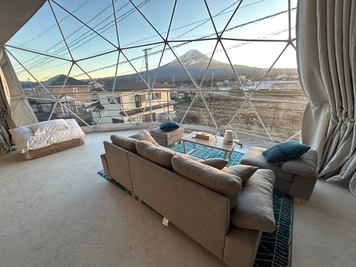 A spacious living area features large panoramic windows, offering views of Mount Fuji. Soft furnishings include a sectional couch and scattered cushions, alongside a low coffee table. Natural light fills the room, enhancing the minimalist design. A second sleeping area is visible in the background.