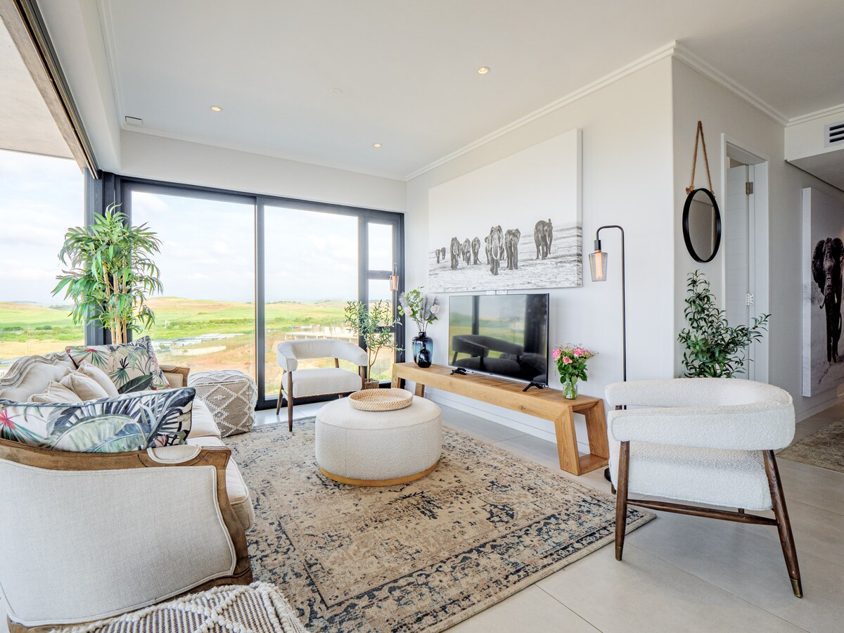 A light-filled lounge area is showcased, featuring a large wall-mounted TV and a wooden console. Soft seating options include a white armchair and a patterned sofa. Expansive glass doors reveal views of the surrounding landscape, complemented by indoor plants and decorative art.