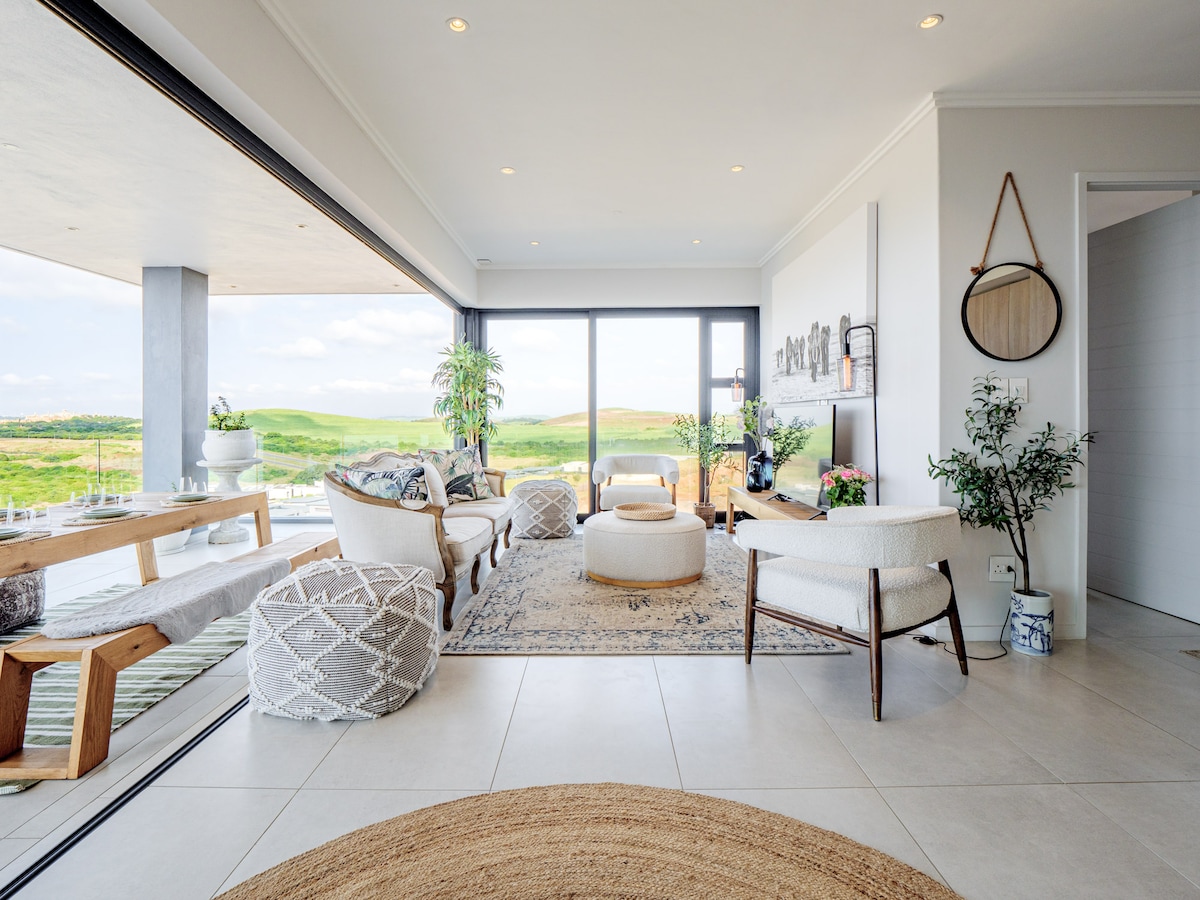 A spacious lounge features large glass doors that open to a breathtaking view of the landscape. Comfortable seating, including patterned chairs and a light-colored ottoman, surrounds a central area rug. A dining space with natural wood accents is visible in the background, enhancing the open layout.