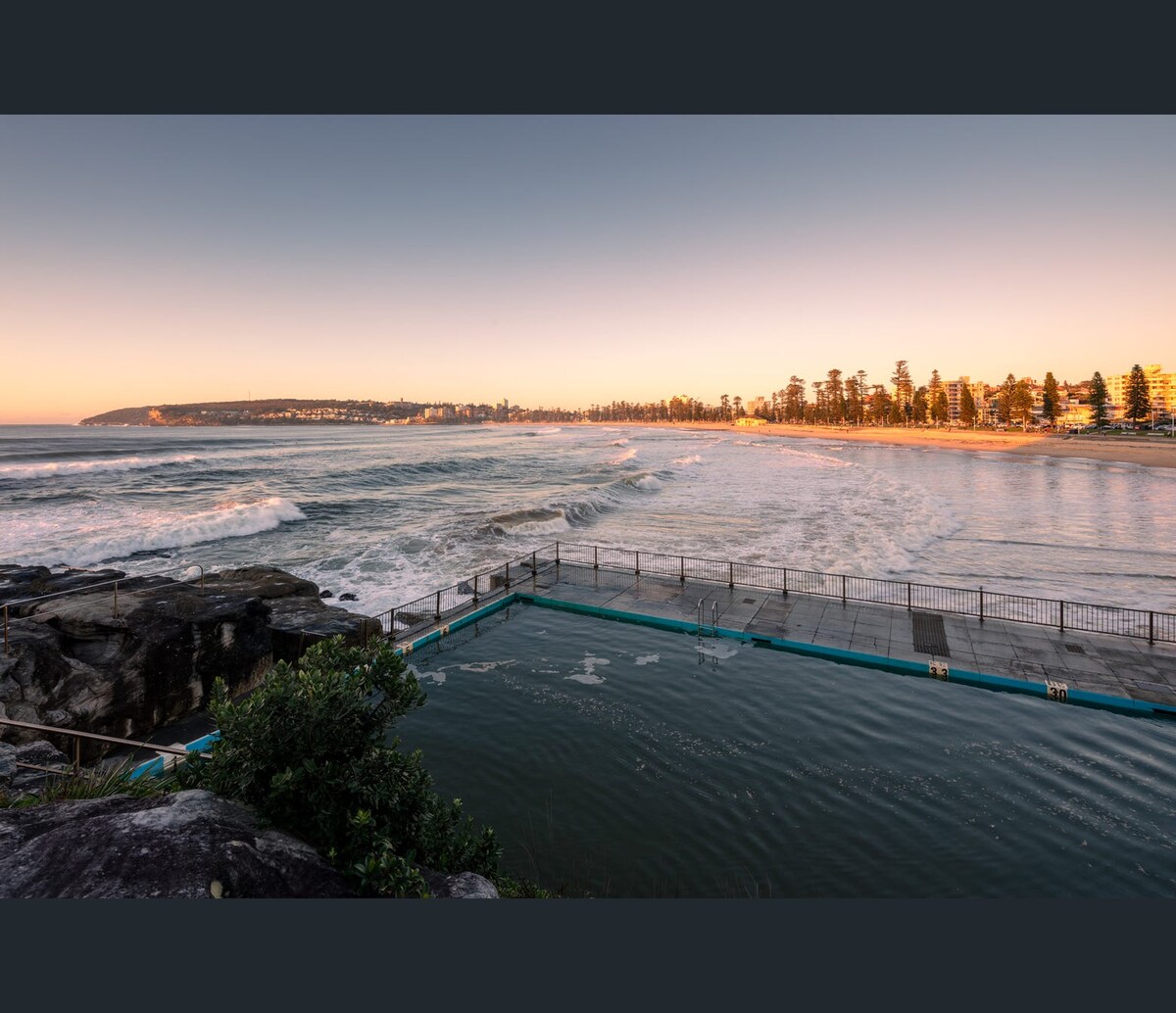 The image captures a serene coastal scene featuring a tidal pool against a backdrop of gentle waves and distant shoreline. The warm colors of the sunset illuminate the water, while rocky formations and greenery frame the foreground, creating a harmonious connection with nature.