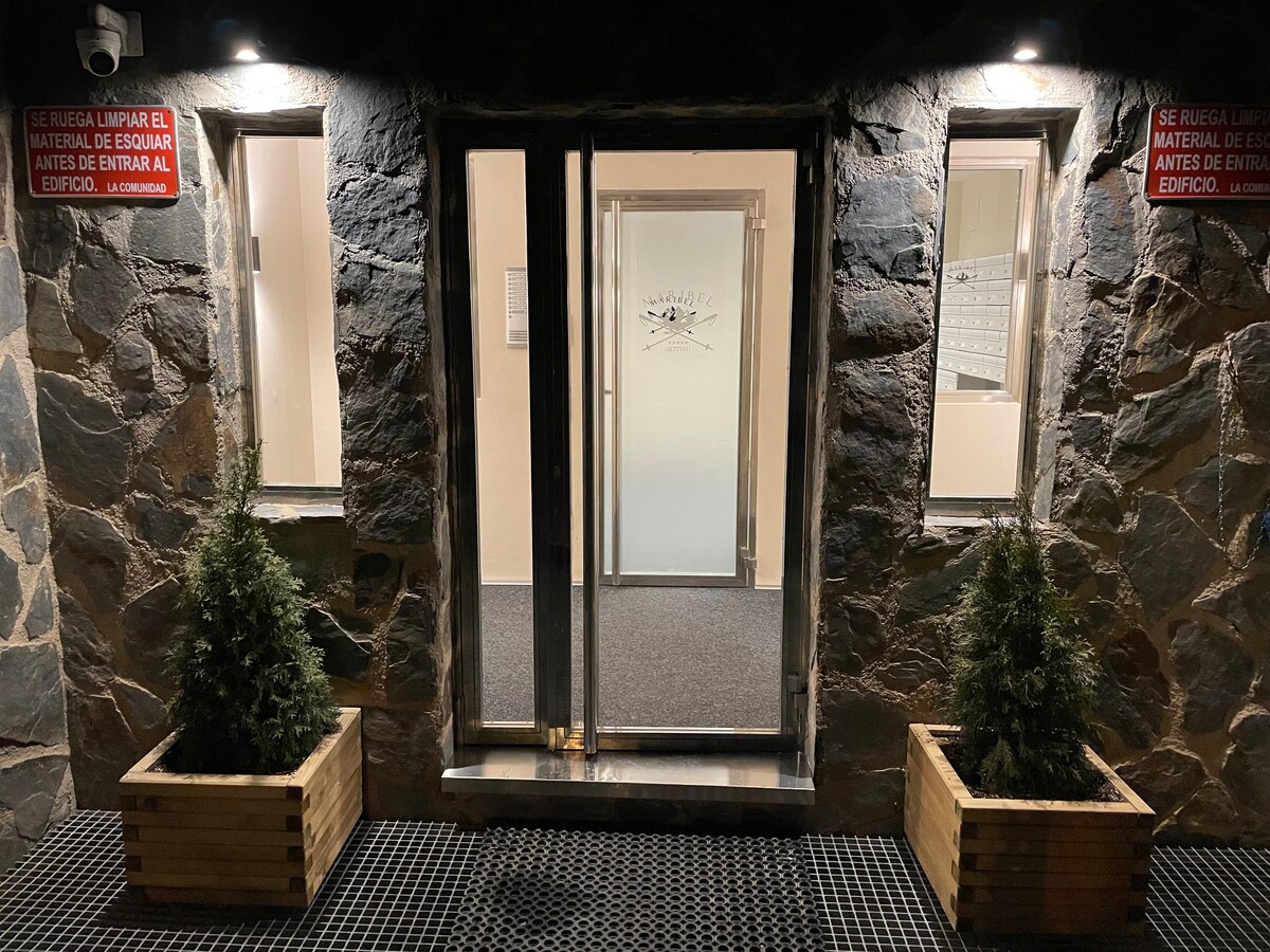 The entrance to the building is framed by natural stone walls, featuring glass double doors that provide access inside. Planters with small evergreen plants are positioned on either side of the entrance. Soft lighting enhances the doorway's visibility during evening hours.