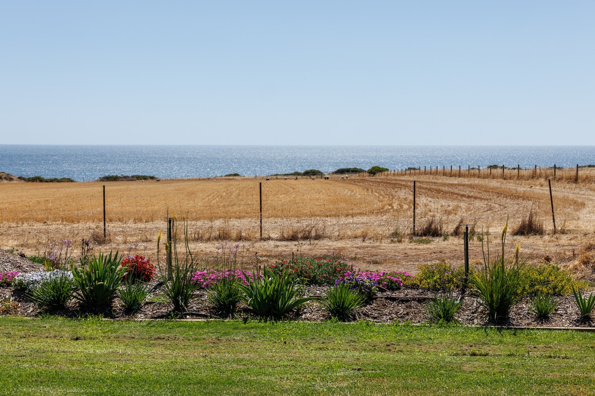 A scenic view of golden coastal farmland meets the blue waters of the ocean. A vibrant flower bed in the foreground features colorful blooms, while a rustic fence divides the landscape. The clear sky enhances the serene atmosphere of this coastal retreat.
