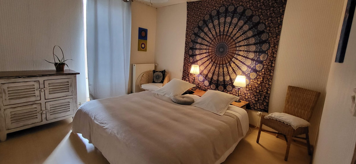A spacious bedroom is presented, featuring a large bed with neutral bedding. Two bedside lamps are positioned on either side, casting a warm glow. A decorative tapestry hangs on the wall, and natural light filters through sheer curtains. A wicker chair and a wooden dresser complete the space.