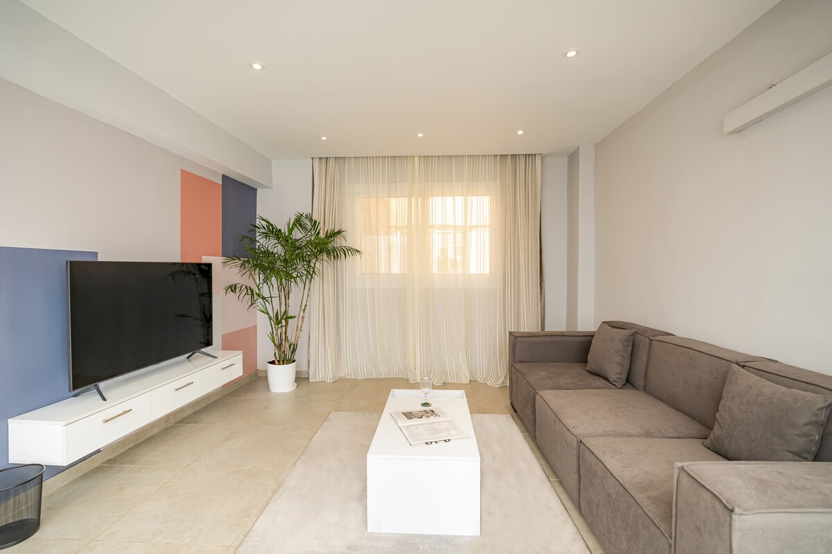 A modern living area features a large, gray sectional sofa positioned opposite a sleek television on a white media console. Natural light filters through sheer curtains, illuminating a minimalist coffee table centered on a soft area rug. A potted plant adds a touch of greenery.