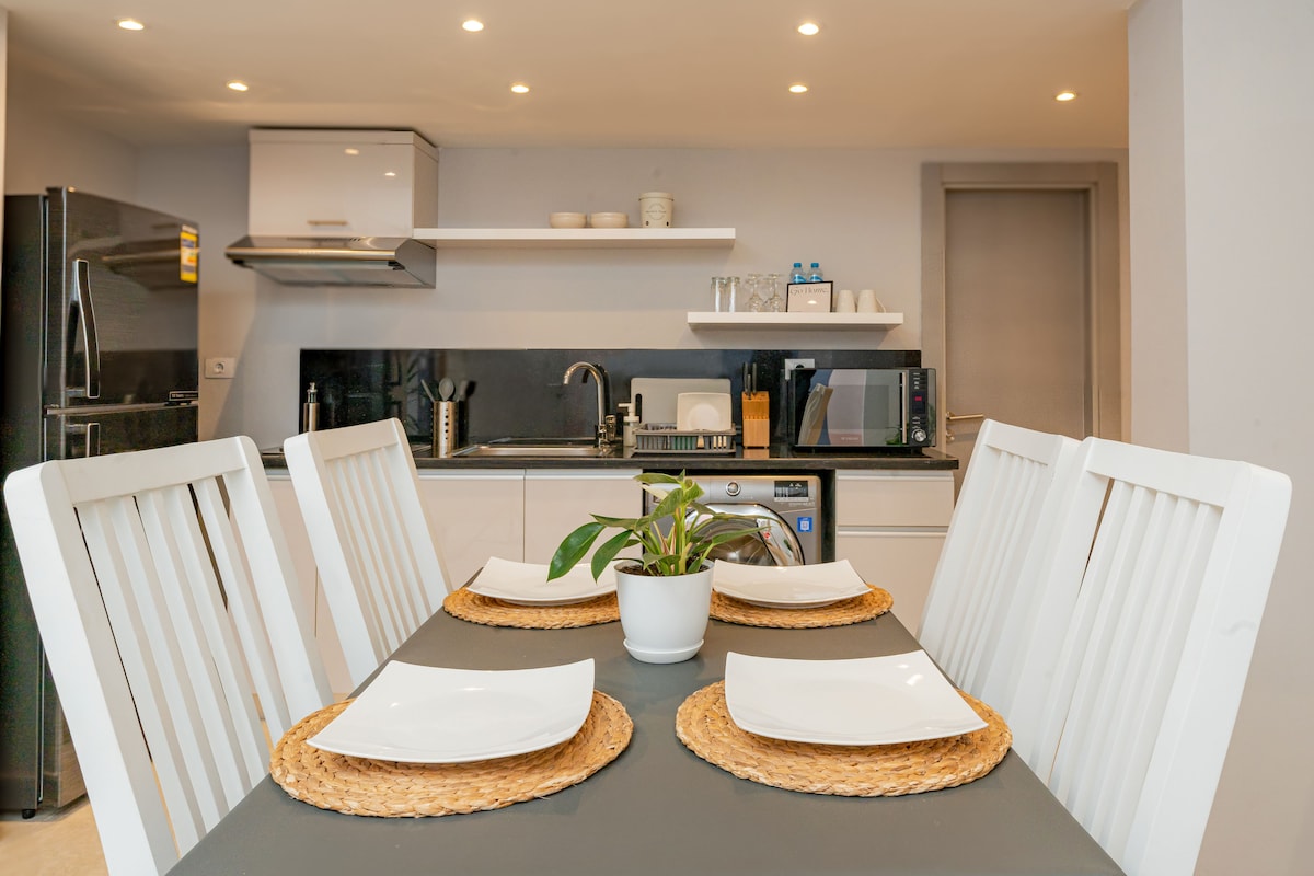 A modern kitchen is showcased with a sleek countertop and organized shelves. A dining table, set for four, features white plates and woven placemats. Appliances, including a microwave and coffee maker, are neatly arranged. A small plant adds a touch of greenery to the setting.