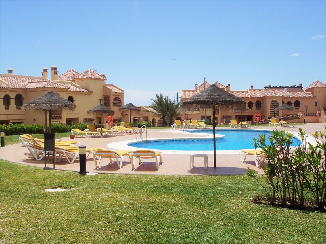 A spacious pool area is surrounded by lounge chairs under straw umbrellas. Well-maintained gardens with green lawns complement the inviting swimming pool, while modern Mediterranean-style buildings frame the scene. Clear blue skies enhance the relaxing atmosphere of this residential community.