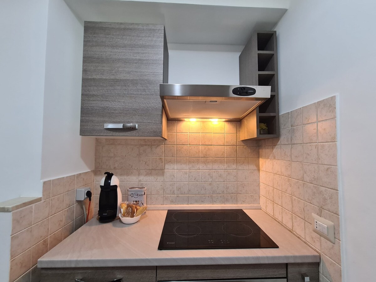 A modern kitchen features a sleek induction cooktop beneath a stainless steel range hood. Light emanates from the hood, highlighting the textured tile backsplash. Cabinets with a warm wood finish provide ample storage above, enhancing the clean, contemporary design.