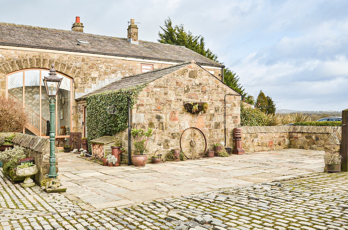 The exterior of the barn conversion features a stone facade with wooden accents, surrounded by landscaped stone paths and planters. A vintage lantern stands beside a paved entrance, leading to a serene outdoor area that conveys a rustic charm complemented by natural greenery.