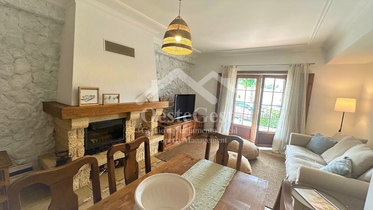 A welcoming living area features a stone fireplace and a large window allowing natural light. A wooden dining table with seating is positioned next to a cozy sofa. Light curtains frame the window, and decorative wall art adds elegance to the space.