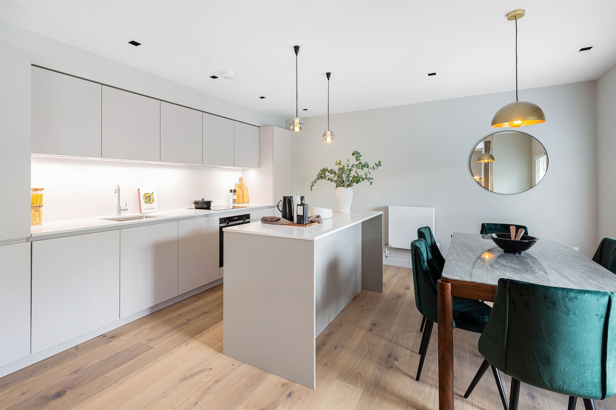 A modern kitchen features sleek cabinetry with integrated appliances. An island with a light-colored countertop sits in the center, complemented by a wooden dining table with green upholstered chairs. Natural light illuminates the space, enhancing the minimalist design.