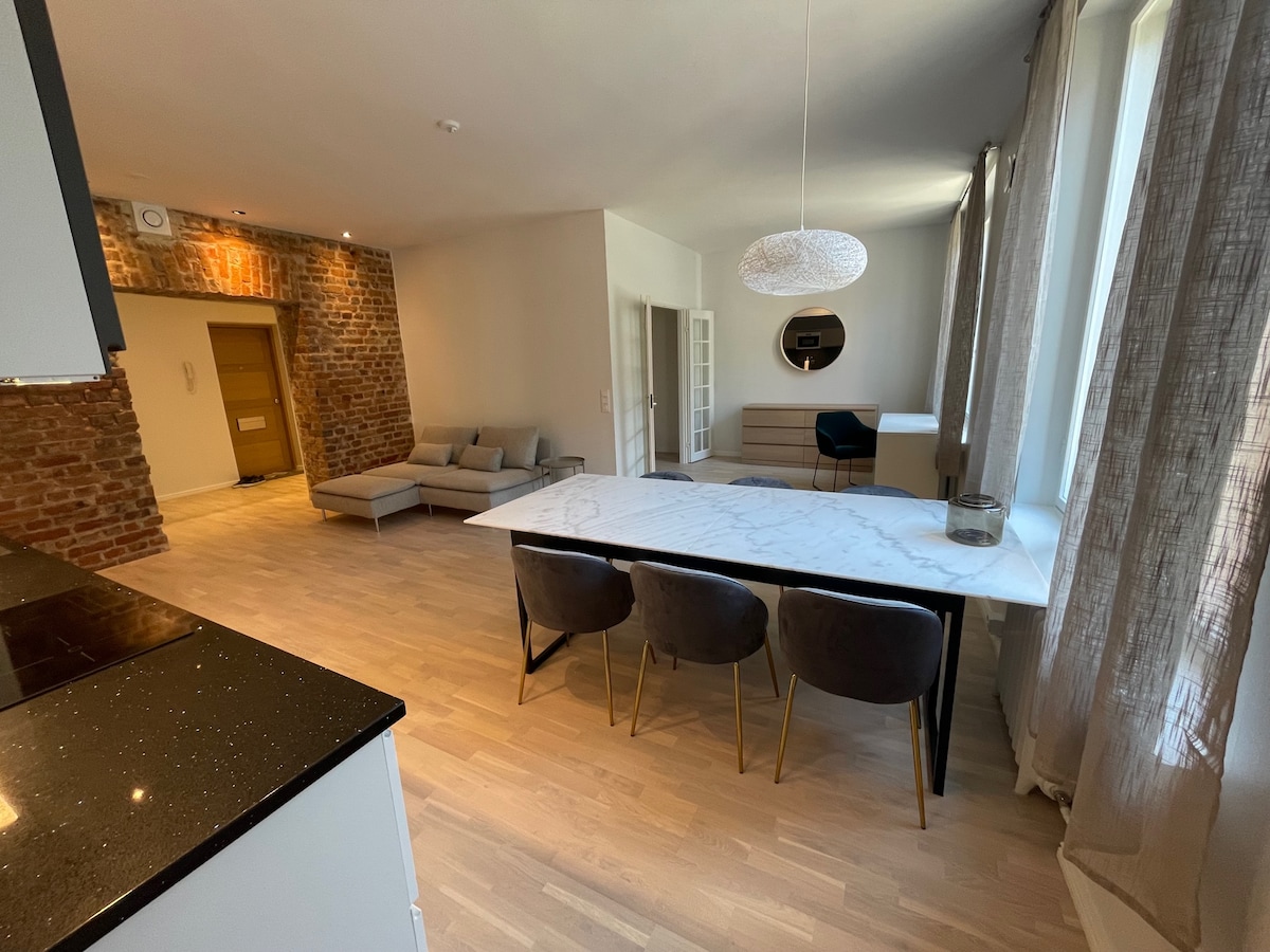 An open living area features a modern dining table surrounded by four upholstered chairs. A sleek sideboard and a circular mirror are positioned against the wall. Natural light filters through large windows, highlighting the brick accent wall and light wood flooring.