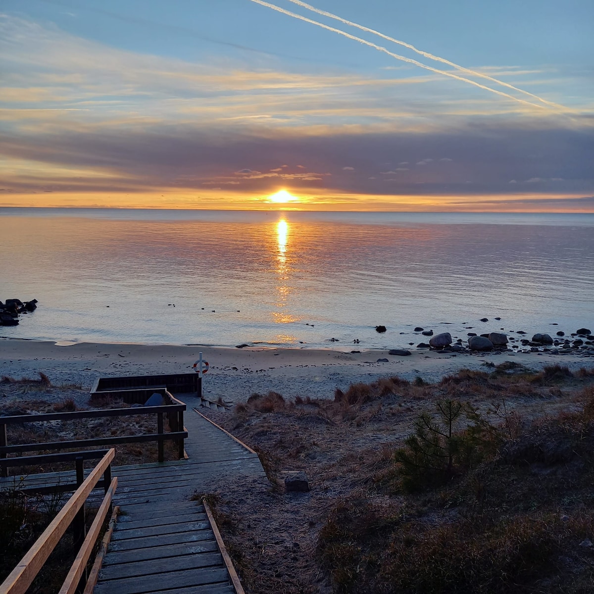 A serene view of the sunrise over a calm sea is captured, with soft colors blending in the sky. A wooden pathway leads down to the beach, framed by gentle sand dunes and scattered rocks along the shoreline.