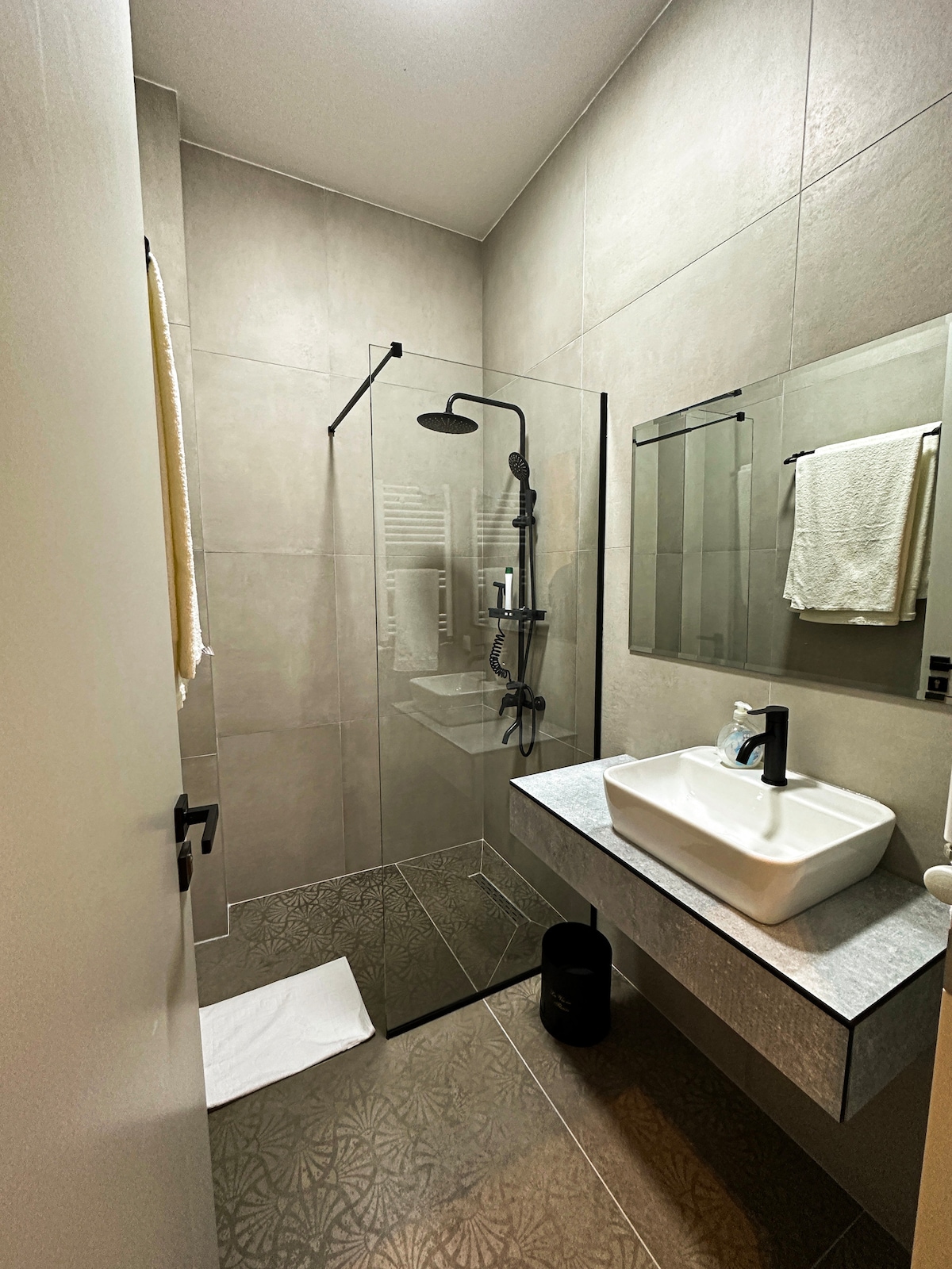 The bathroom features modern finishes with textured flooring and neutral tile walls. A glass shower enclosure is complemented by a sleek showerhead. A contemporary sink rests on a floating vanity, and a mirror reflects the minimalistic design, including neatly arranged toiletries and a towel.