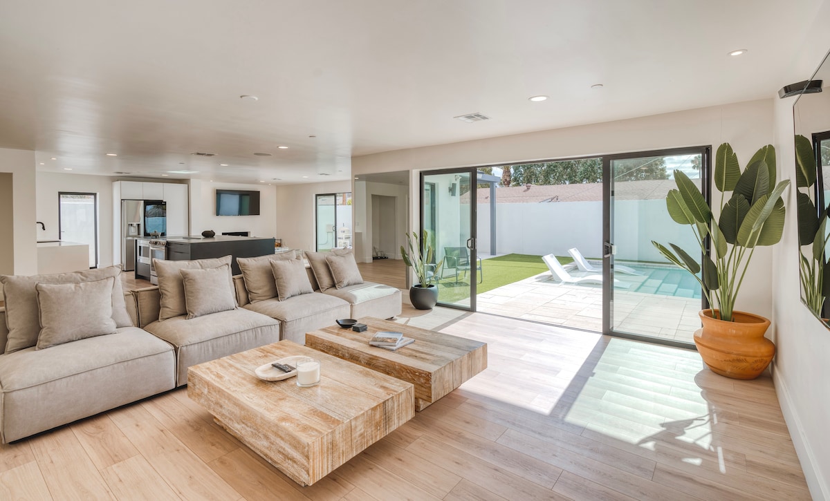 A spacious living area is filled with natural light from large glass doors that open to the backyard. A comfortable sectional sofa is complemented by two wooden coffee tables. Lush indoor plants are placed in a terracotta pot, while a view of the pool and lounge chairs is visible beyond.