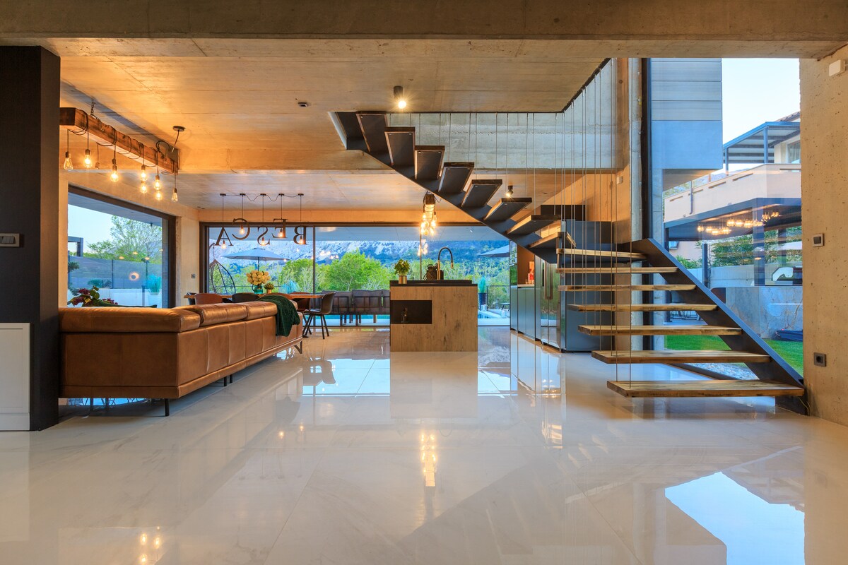 A modern living area features polished marble flooring and large glass windows that provide natural light. An elegant staircase made of wood and metal leads to the upper level. A comfortable leather sofa and a dining area are visible, enhancing the spacious layout.
