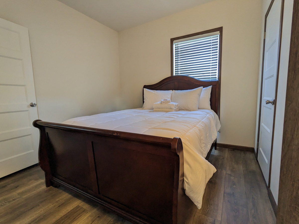 A private sleeping area features a spacious bed with crisp white linens and neatly arranged towels. Natural light filters through a window with horizontal blinds, illuminating warm-toned wood flooring. Two doors, one leading to the room and another to the neighboring space, are visible.