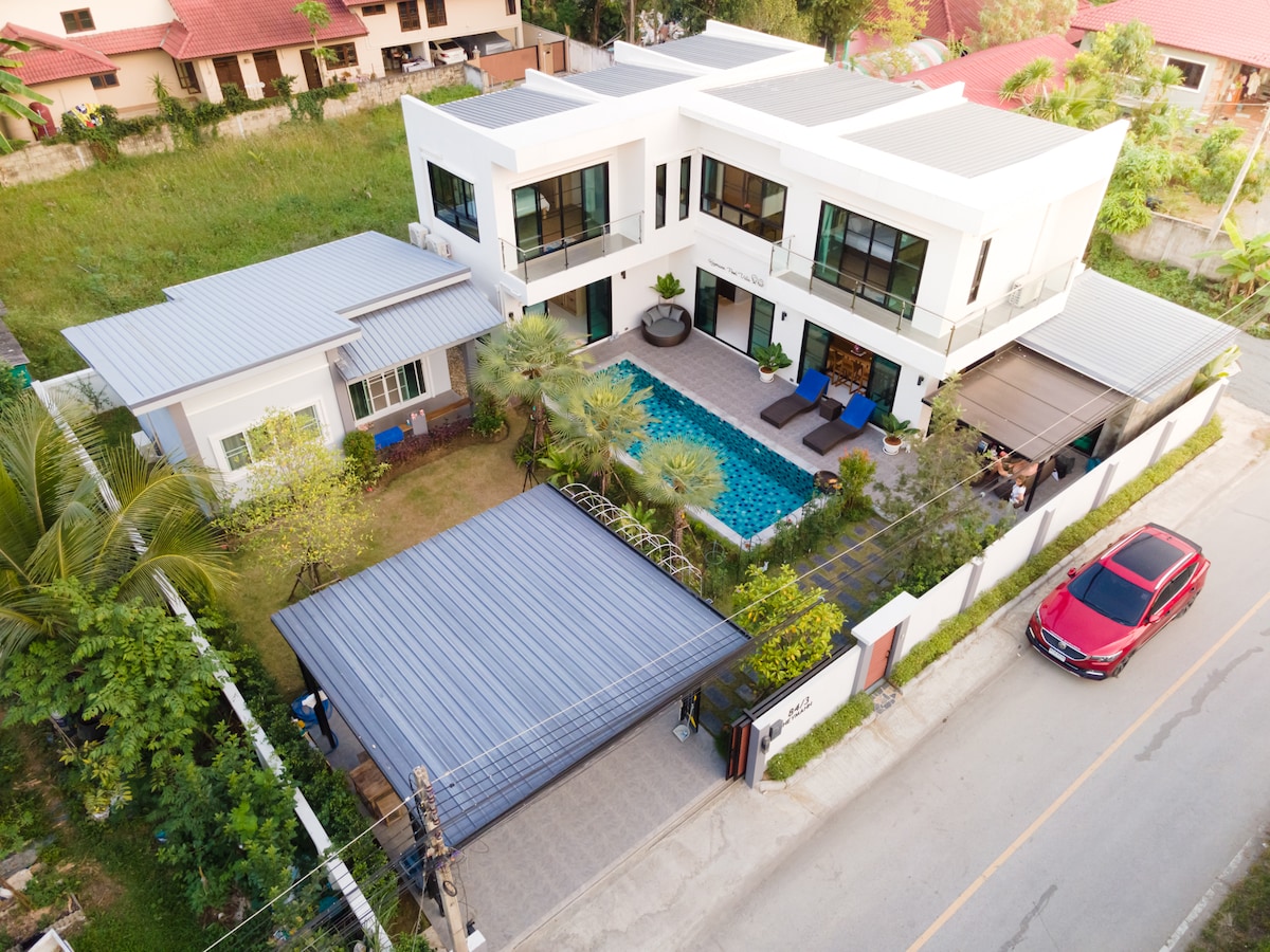 The villa's modern architecture is showcased from an aerial view, highlighting a lush green yard surrounding a rectangular saltwater pool. Relaxation areas with lounge chairs are positioned near the water. The two-story layout and surrounding foliage contribute to the overall spaciousness of the property.