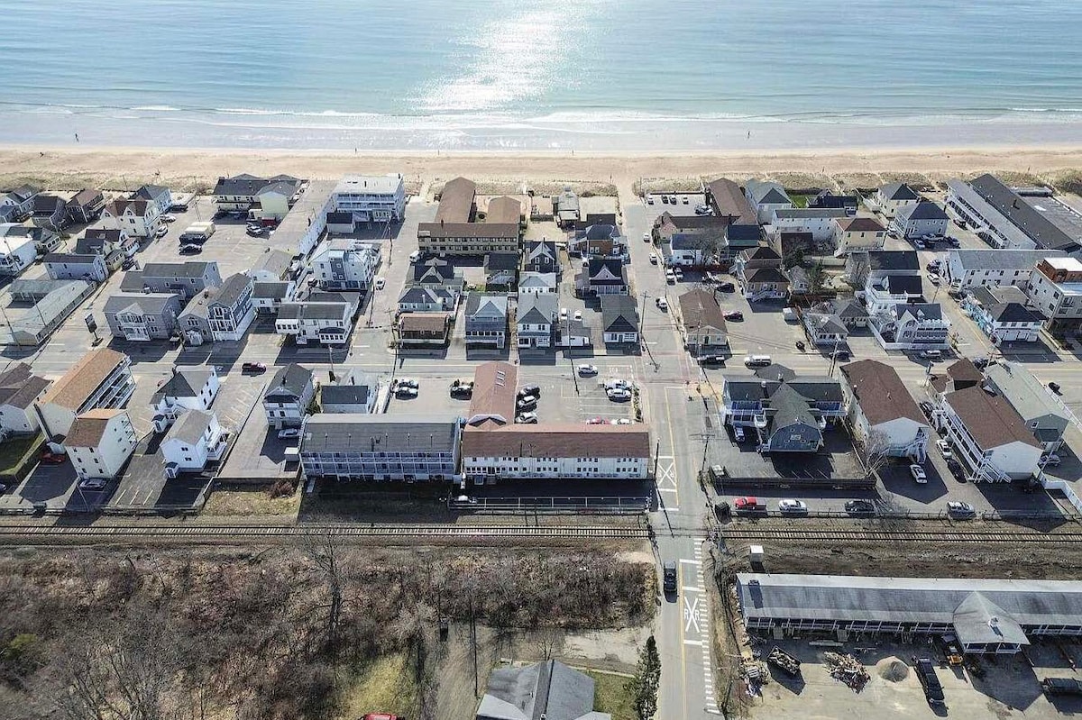 An aerial view captures a coastal community, displaying a grid of residential and commercial buildings adjacent to the ocean. The sandy beach stretches along the water's edge, with parking lots and streets visible. A train track runs parallel to the area, connecting various locations.