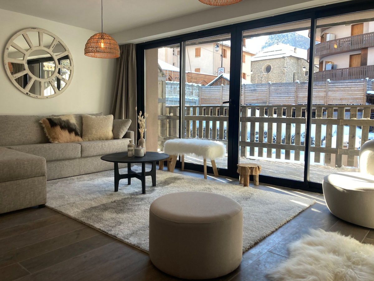 A light-filled living area features a plush grey sofa and a circular coffee table. Two decorative stools and a round pouf are arranged on a soft area rug. Large glass doors open to a private terrace, providing views of the surrounding village.