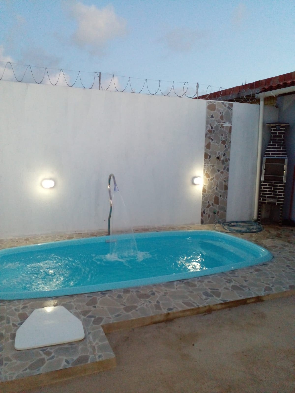 A small pool is seen, surrounded by neutral-colored stone tiles. Soft lighting from wall-mounted fixtures illuminates the area, highlighting the water's clarity. A minimalist shower fixture is positioned at one end, adding functionality to the space.