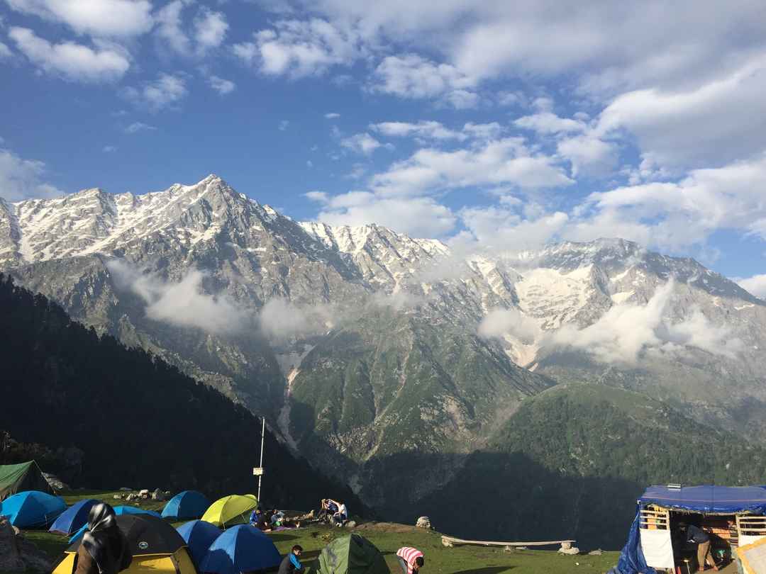 Breathtaking mountain scenery is captured with snow-capped peaks rising majestically under a bright sky. Colorful tents are arranged on a grassy area in the foreground, with soft clouds drifting above the landscape, creating a serene outdoor atmosphere.