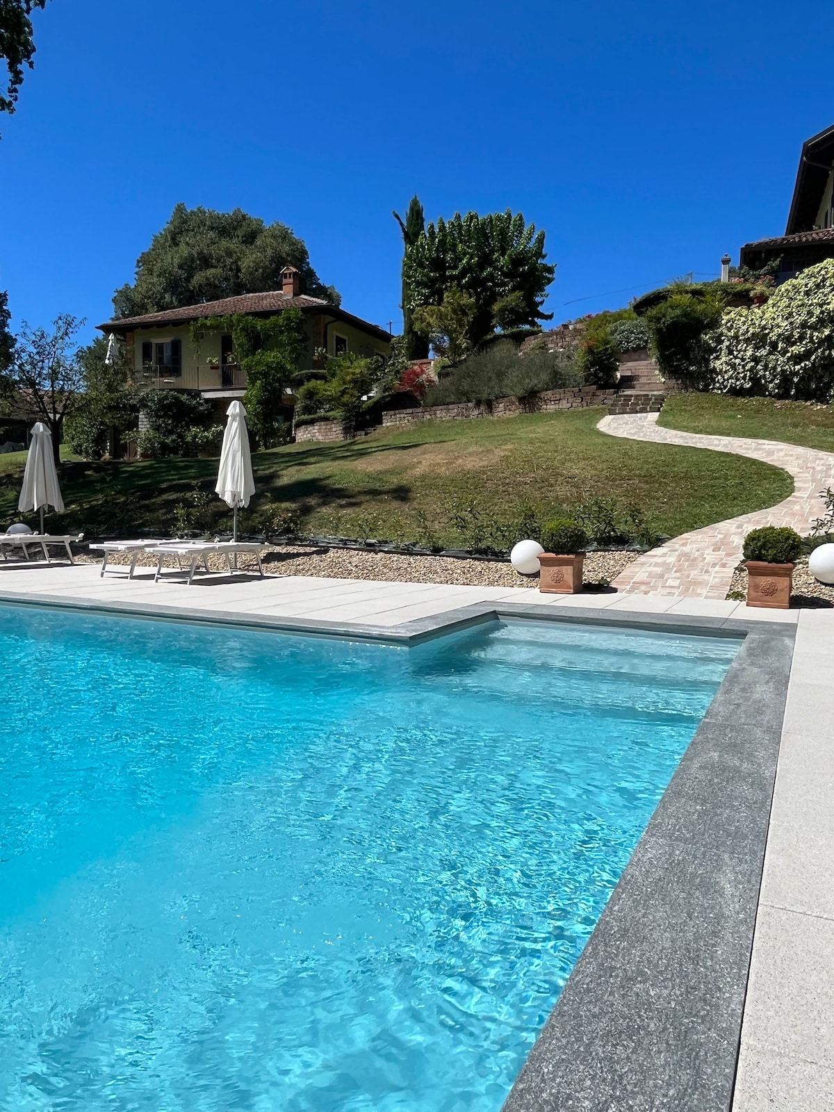 A clear blue swimming pool is framed by a stone terrace. Sun loungers are arranged nearby, and white umbrellas provide shade. A winding path leads to a lush green hillside, where a charming house and trees are visible.