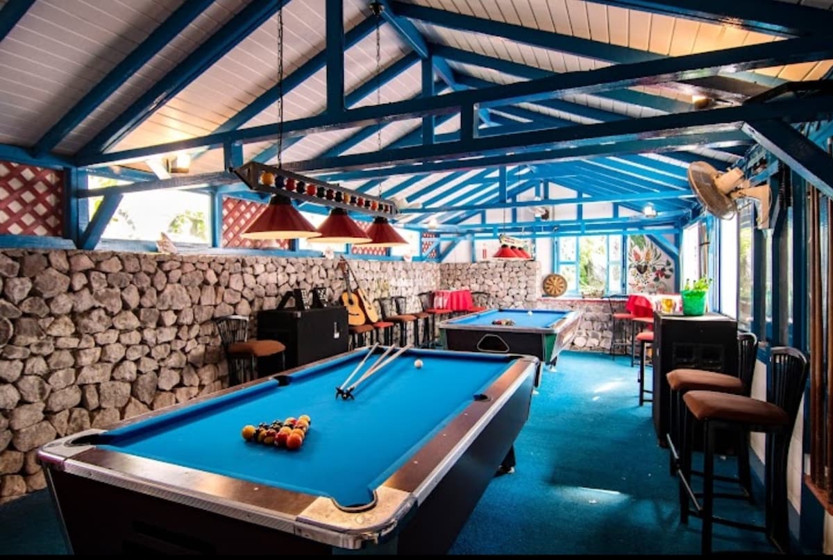 A bright recreational area is highlighted by blue overhead beams and walls adorned with stone. Two pool tables are featured, complemented by overhead lighting. Seating is available at high tables, with a cozy corner showcasing musical instruments and a large window allowing natural light.