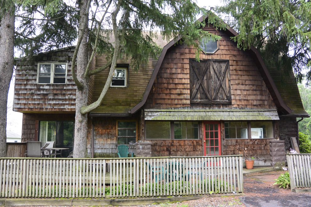 The exterior of a rustic house features a mix of wood siding and a gabled roof. A prominent cross-buck door design is visible on the upper portion. A white picket fence enclosures the yard, while trees provide shade and privacy around the property.