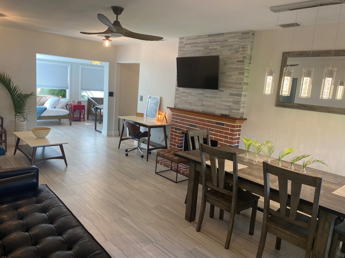 A spacious living area features a comfortable sofa and a large dining table with seating for seven. A mounted television is positioned above a stylish brick fireplace. Natural light filters through the adjacent Florida room, where daybeds are visible, enhancing the open-concept feel.
