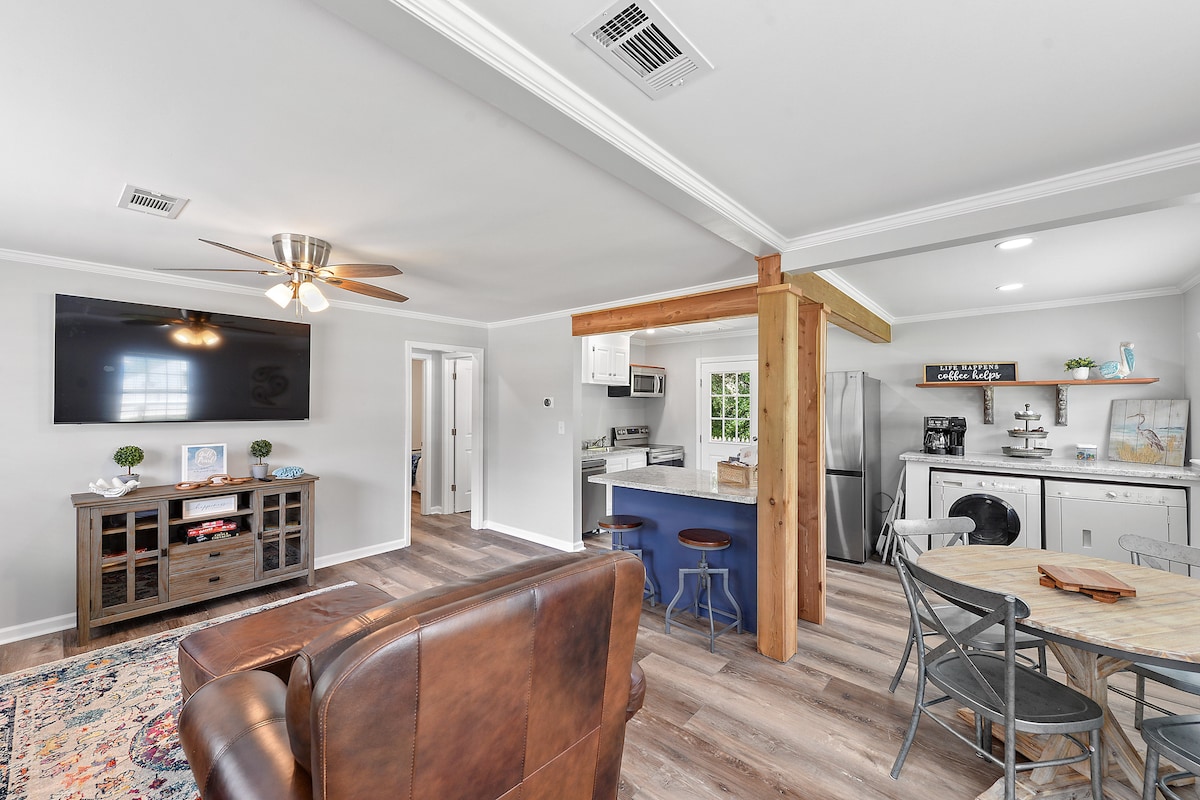 The open living area features a comfortable leather sofa facing a large flat-screen TV mounted on the wall. A dining table for four is situated nearby, with an adjoining kitchen equipped with modern appliances and a rustic wood post dividing the spaces.